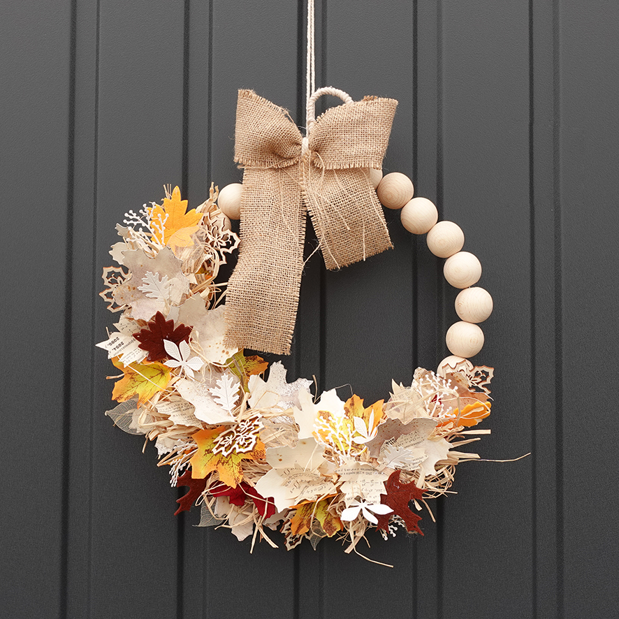 Jesenný venček na dvere z papiera, rafie a drevených korálok je zavesený na tmavošedej bráne.